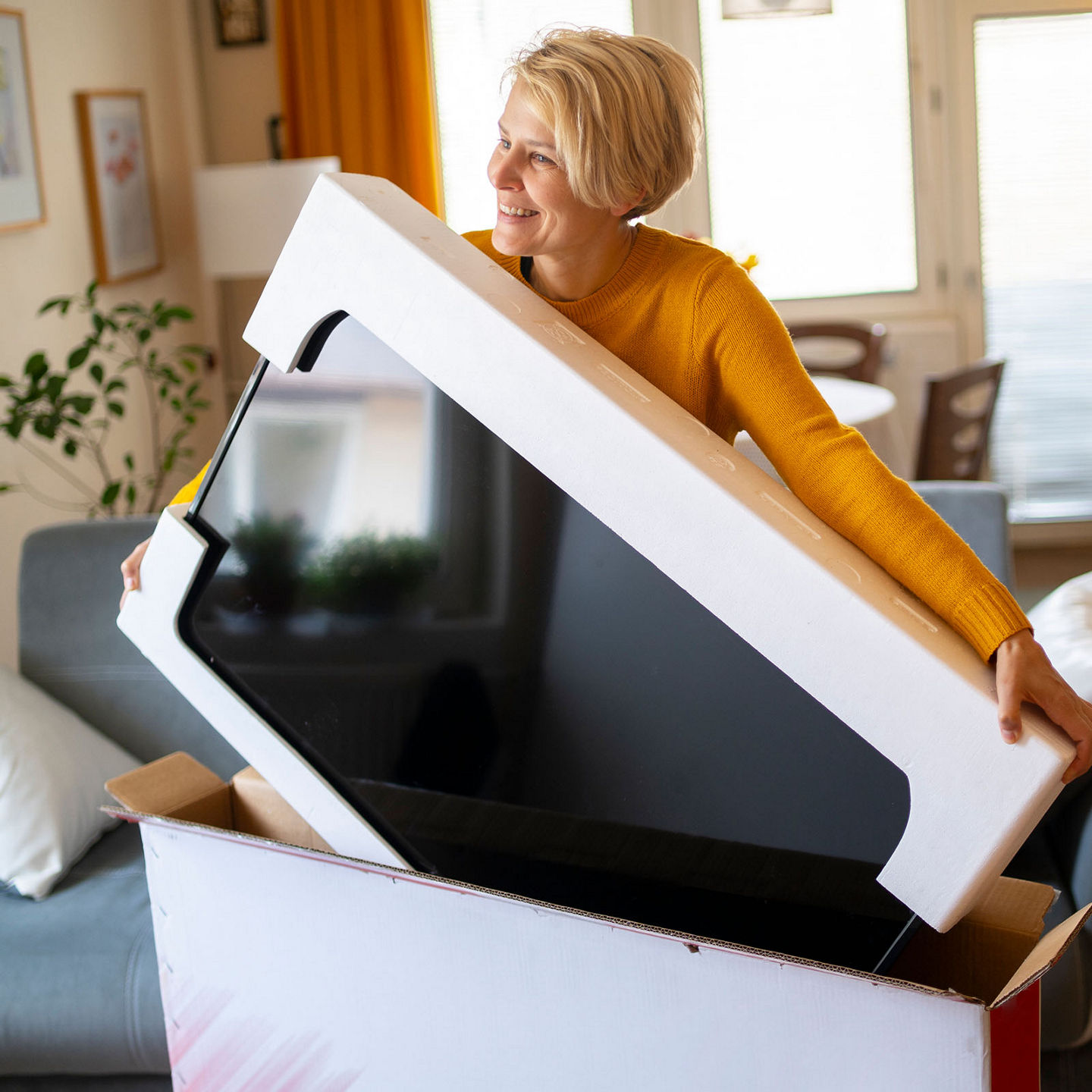 Eine Frau packt hocherfreut einen neuen Fernseher aus dem Versandkarton aus