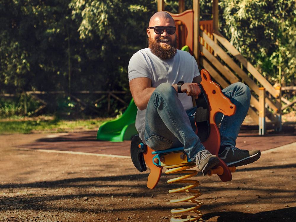 Ein Mann mit Sonnenbrille reitet vergnügt auf einem Spielplatz auf einem Schaukelpferd