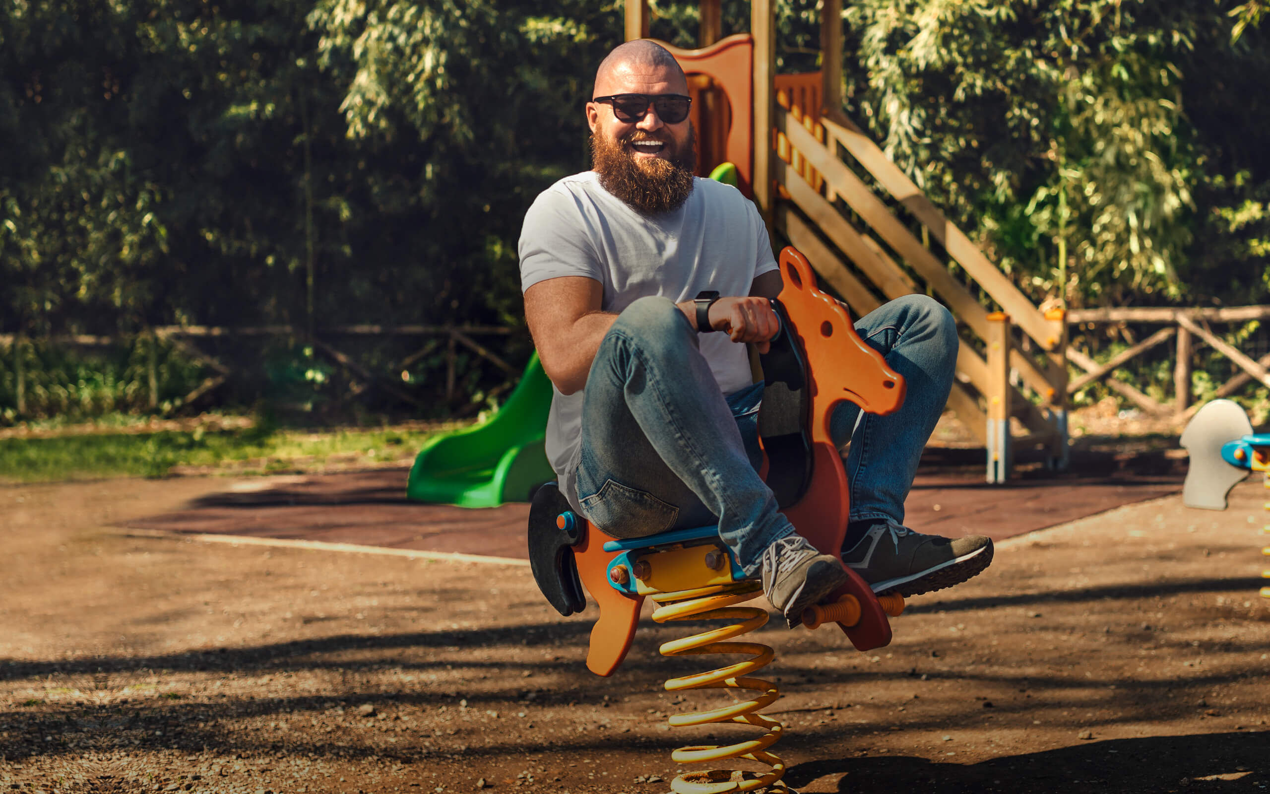 Ein Mann mit Sonnenbrille reitet vergnügt auf einem Spielplatz auf einem Schaukelpferd