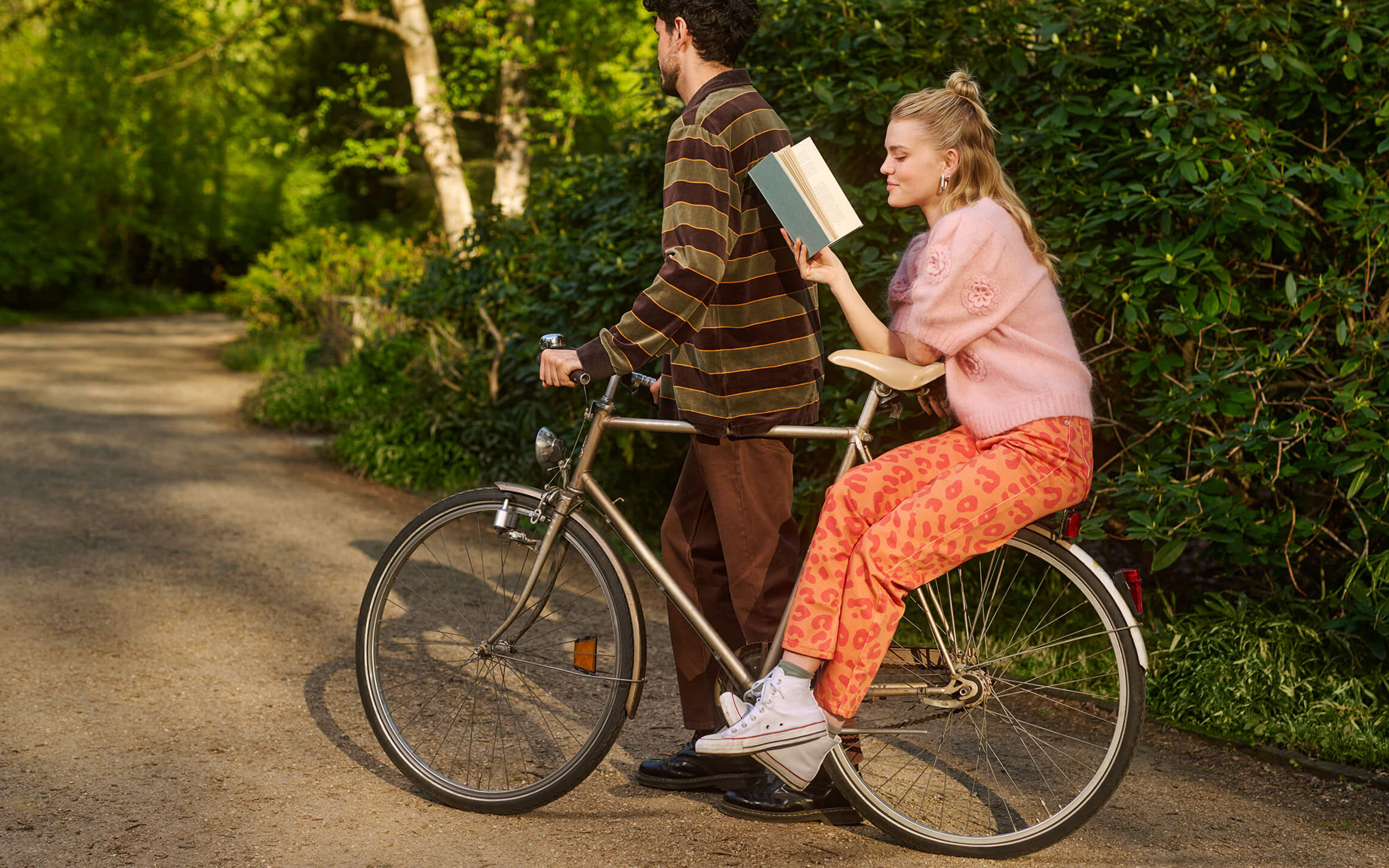 Eine Mann schiebt ein Fahrrad, eine Frau sitzt auf dem Gepäckträger und liest ein Buch