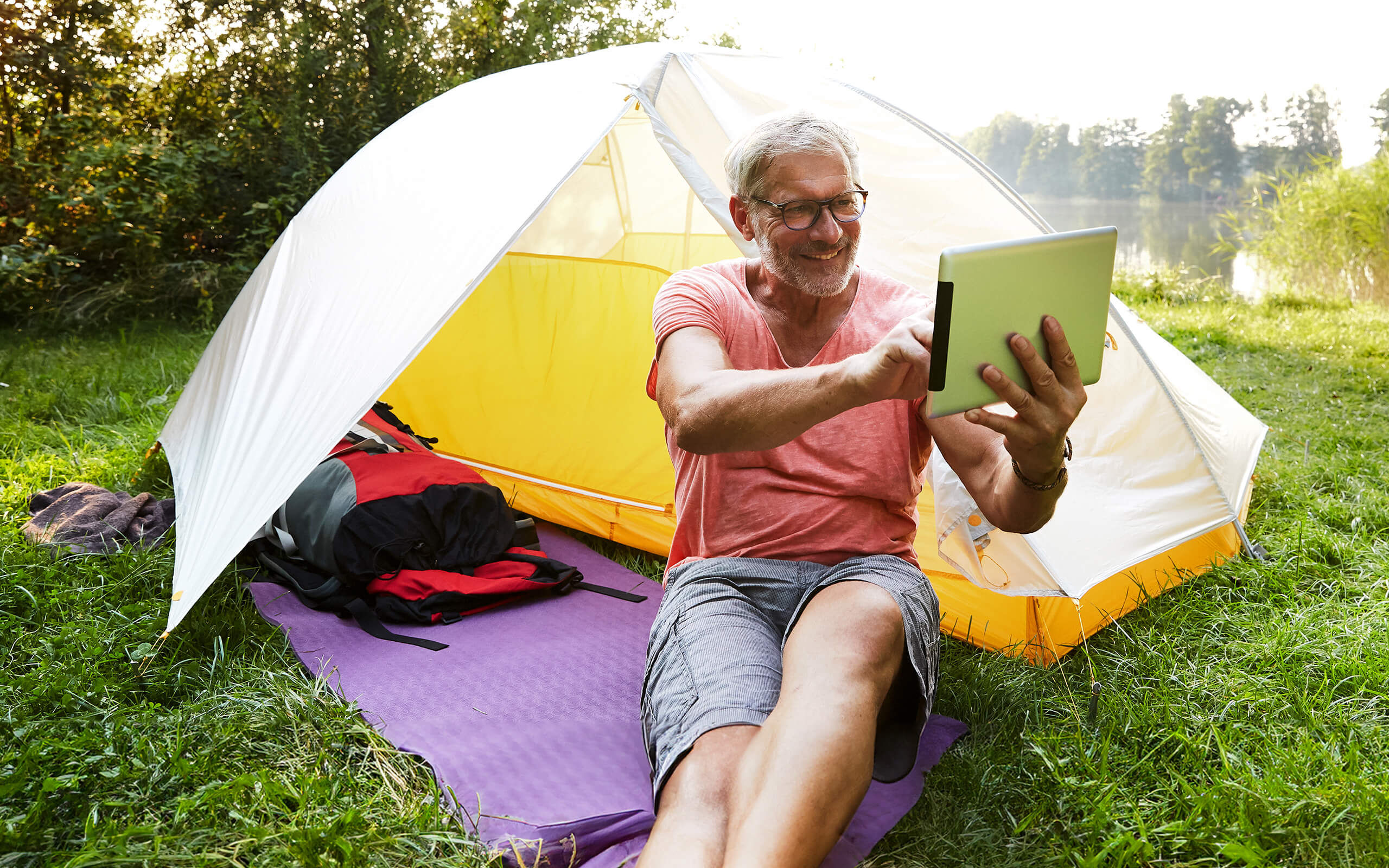 Ein Mann sitzt vor einem kleinen Zelt an einem See und hält ein Tablet in der Hand