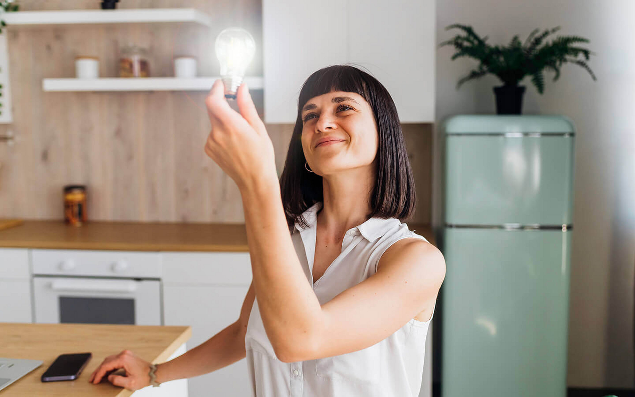 Eine Frau sitzt am Küchentisch und hält eine leuchtende Glühlampe in der Hand