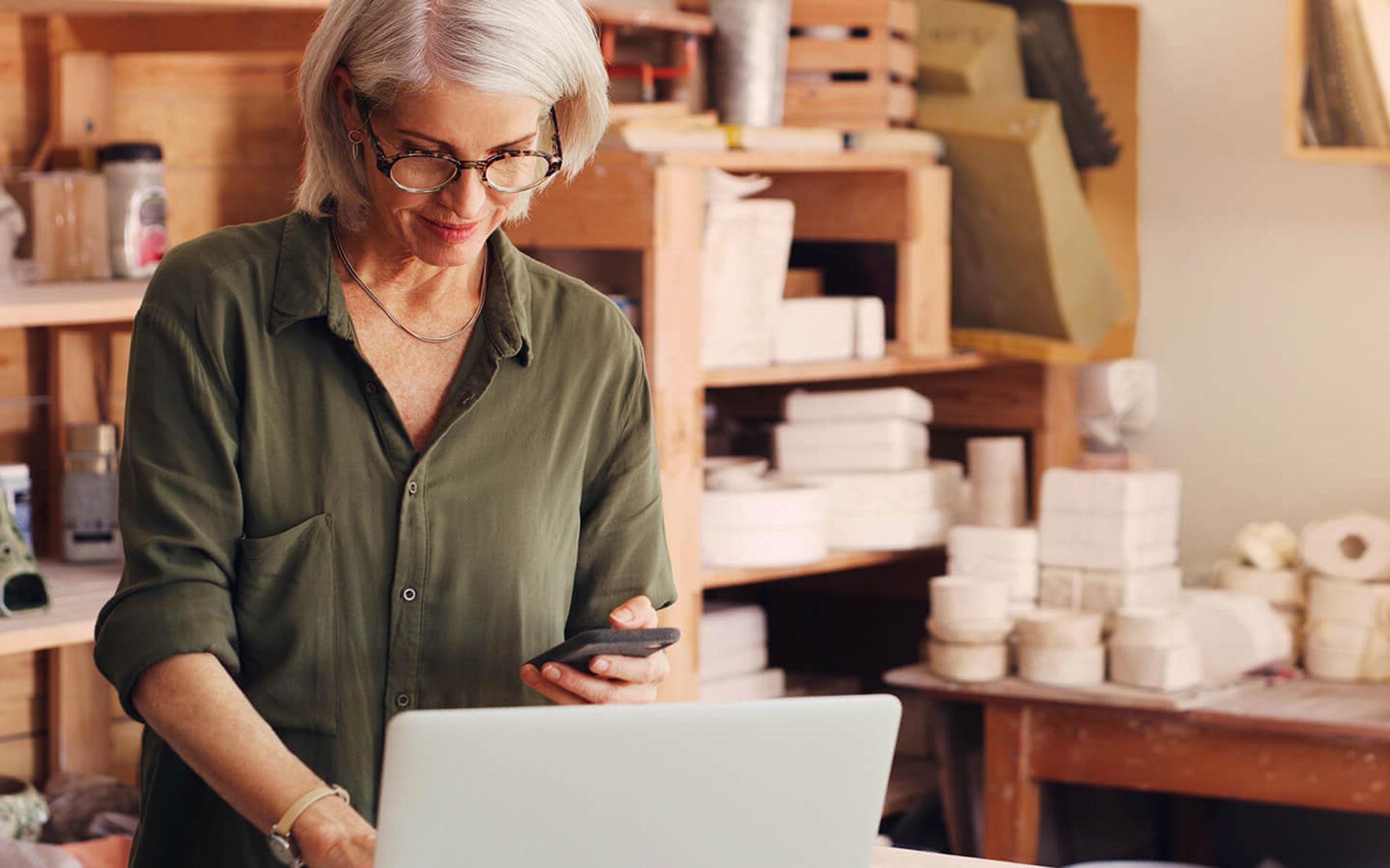 Frau steht in einer Kreativwerkstatt und bedient Laptop und Smartphone gleichzeitig