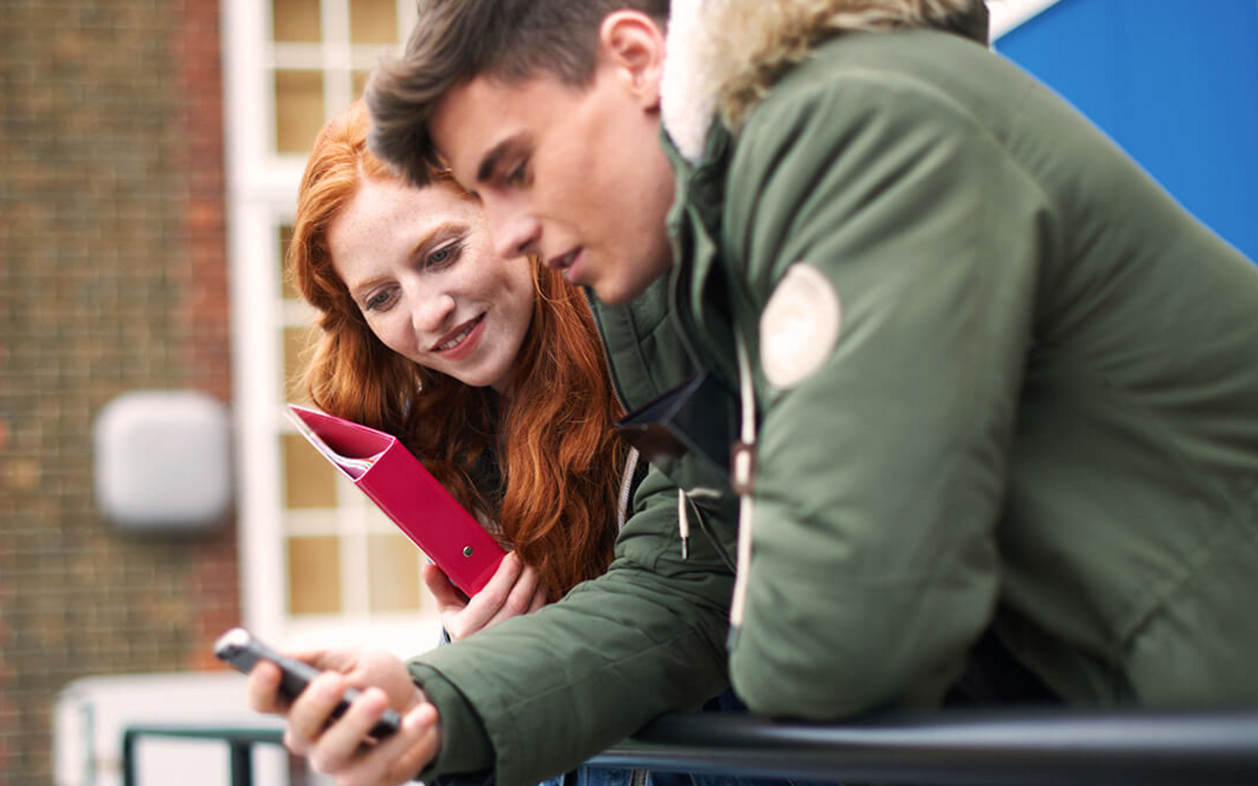 Ein junges Paar lehnt auf einem Geländer und schaut gemeinsam auf ein Smartphone