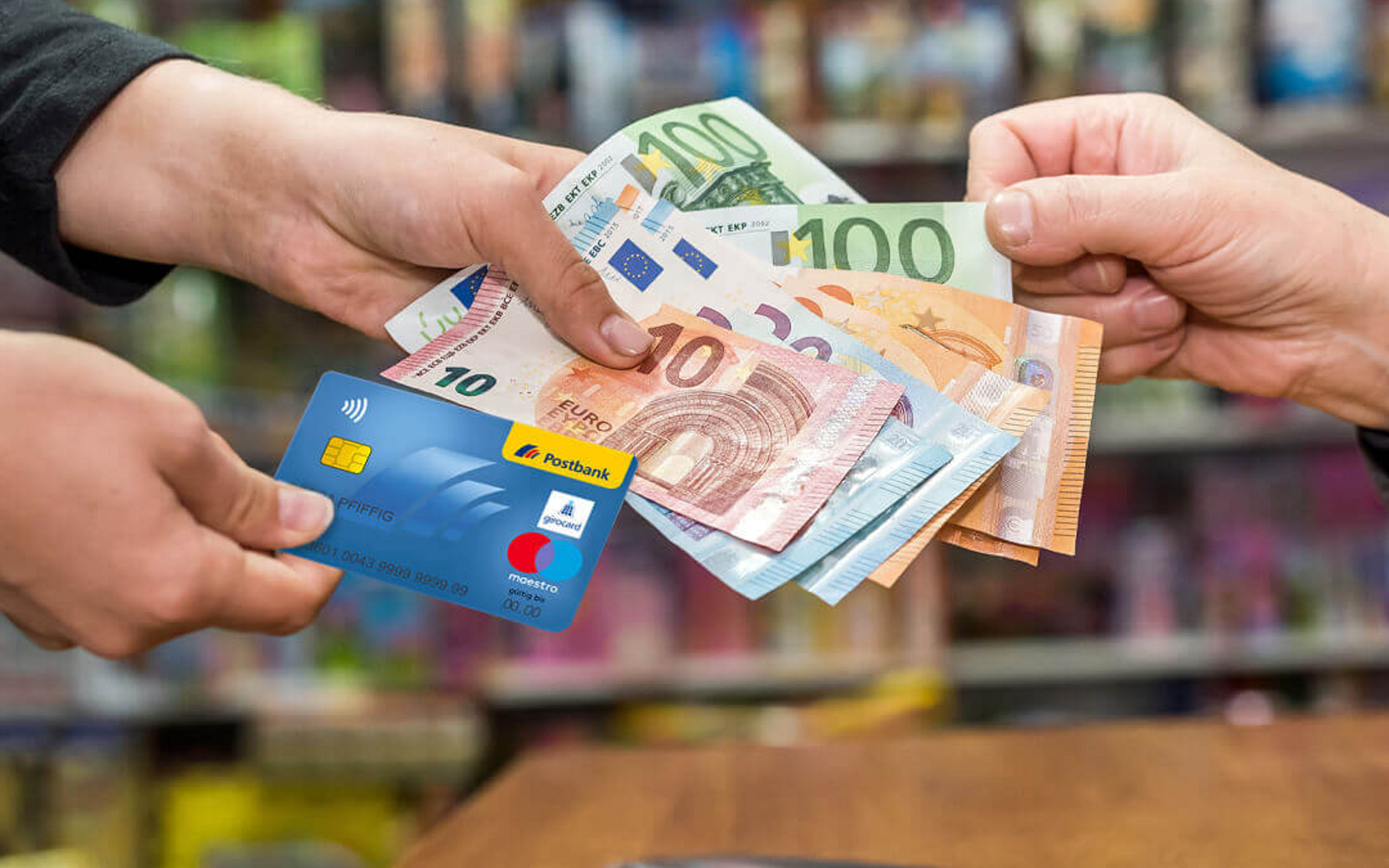 Hände von zwei Personen halten eine EC-Karte der Postbank und Bargeld