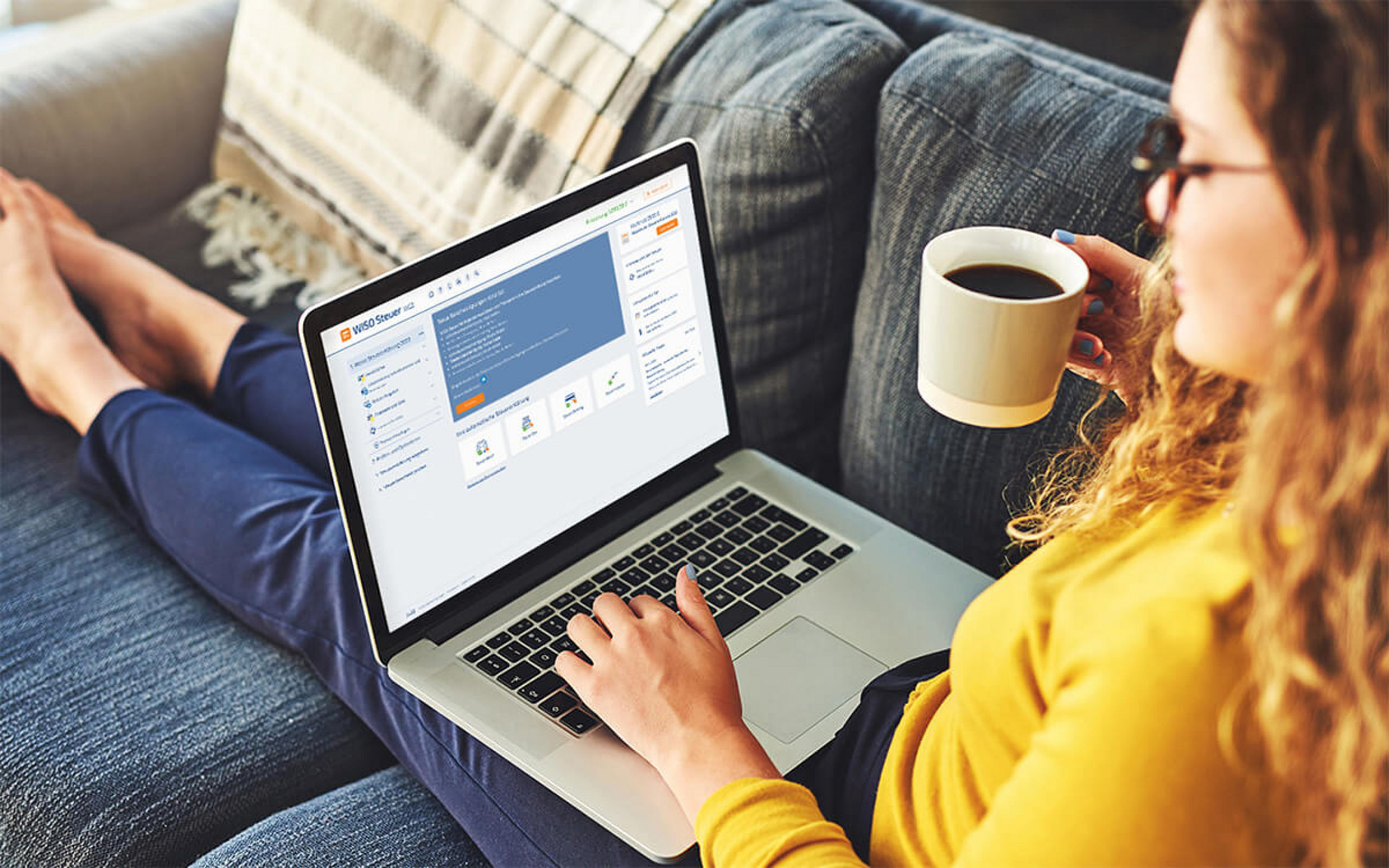 Eine junge Frau sitzt mit einem Kaffee auf der Couch und beliest sich über Finanzen auf Ihrem Laptop