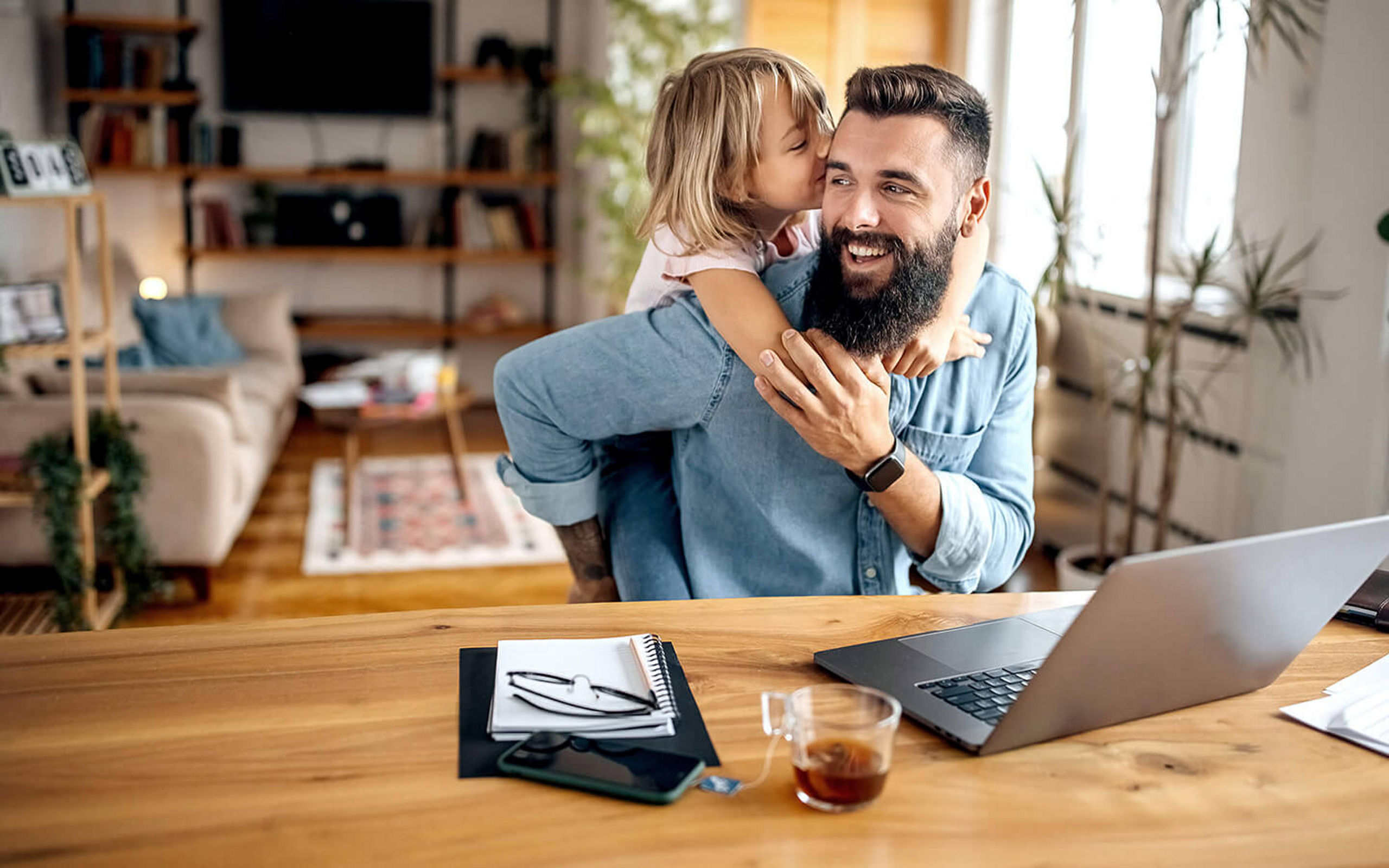 Ein Vater sitzt am Laptop und wird von seiner Tochter umarmt