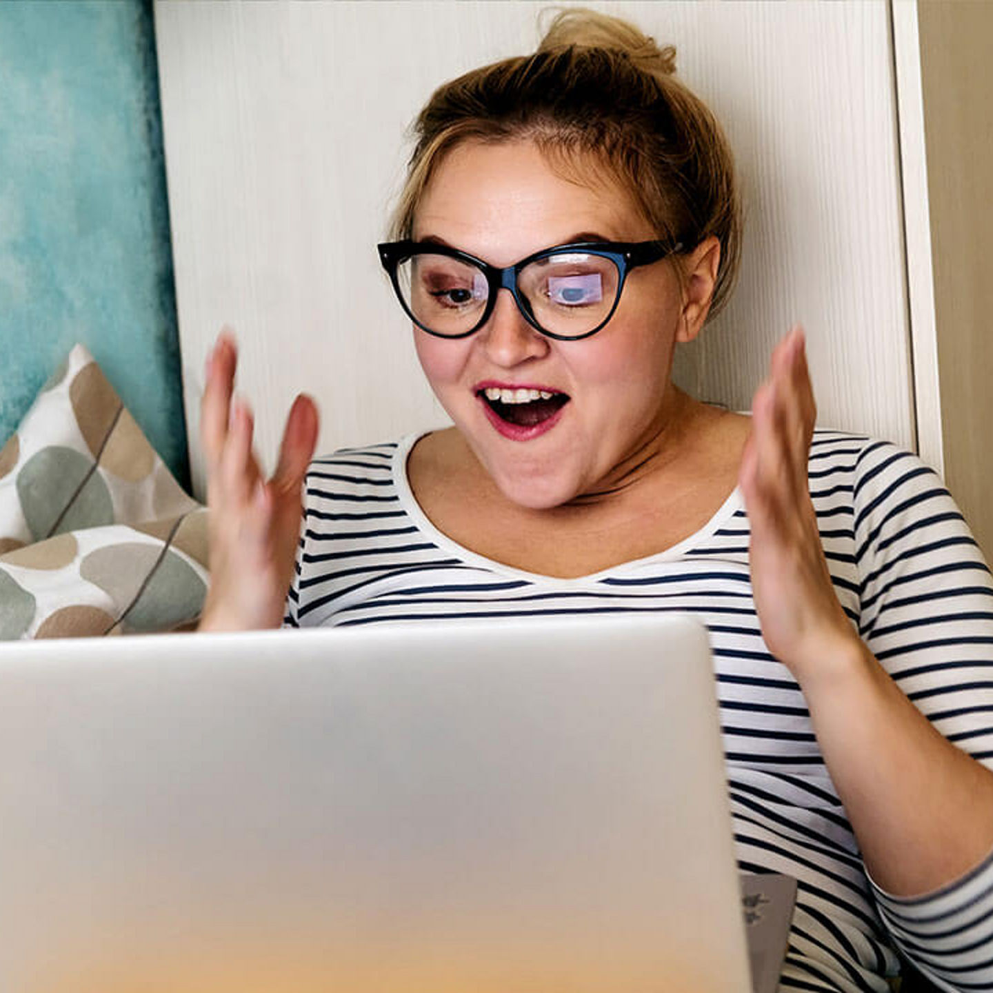 Eine Frau sitzt gemütlich auf einer Couch und schaut hoch erfreut auf ihren Laptop