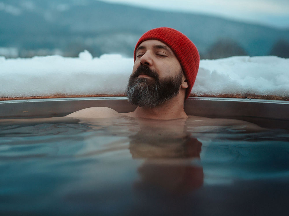 Ein Mann mit roter Mütze entspannt in einem dampfenden Wasserbecken im Freien, umgeben von Schnee