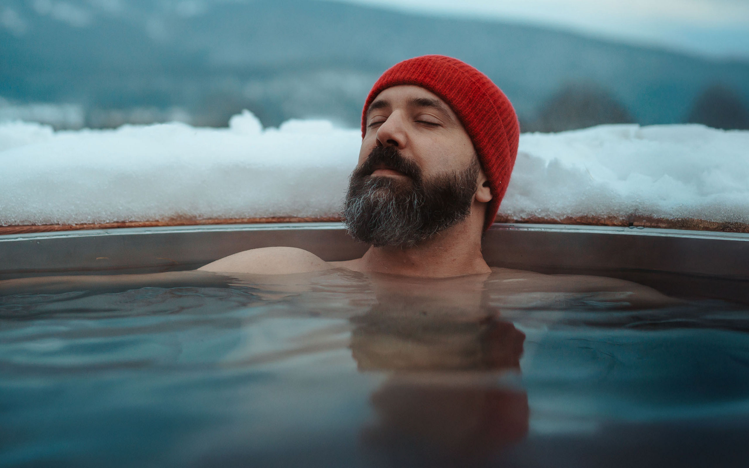 Ein Mann mit roter Mütze entspannt in einem dampfenden Wasserbecken im Freien, umgeben von Schnee