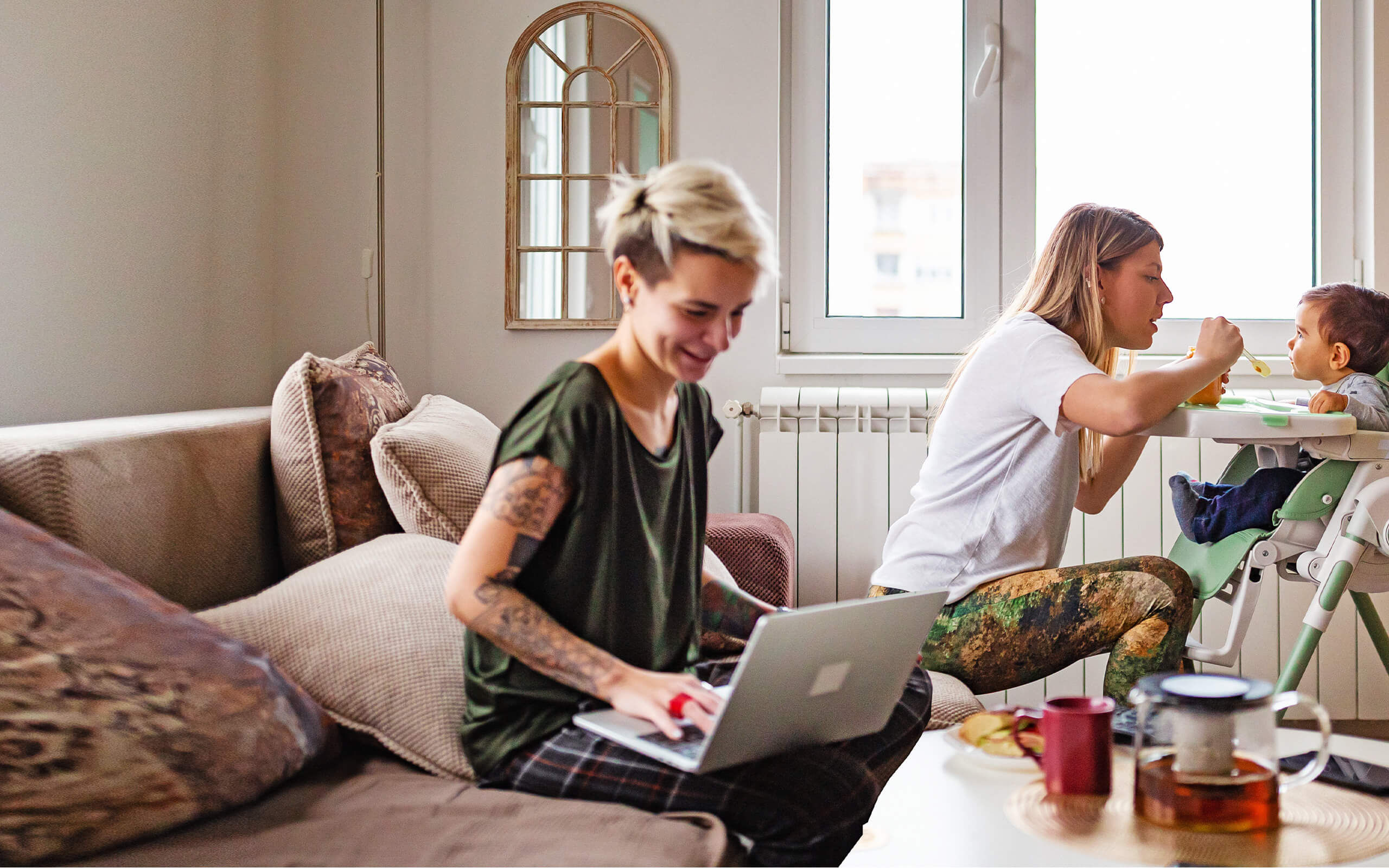 Eine Frau sitzt an einem Laptop, eine andere Frau füttert ein kleines Kind