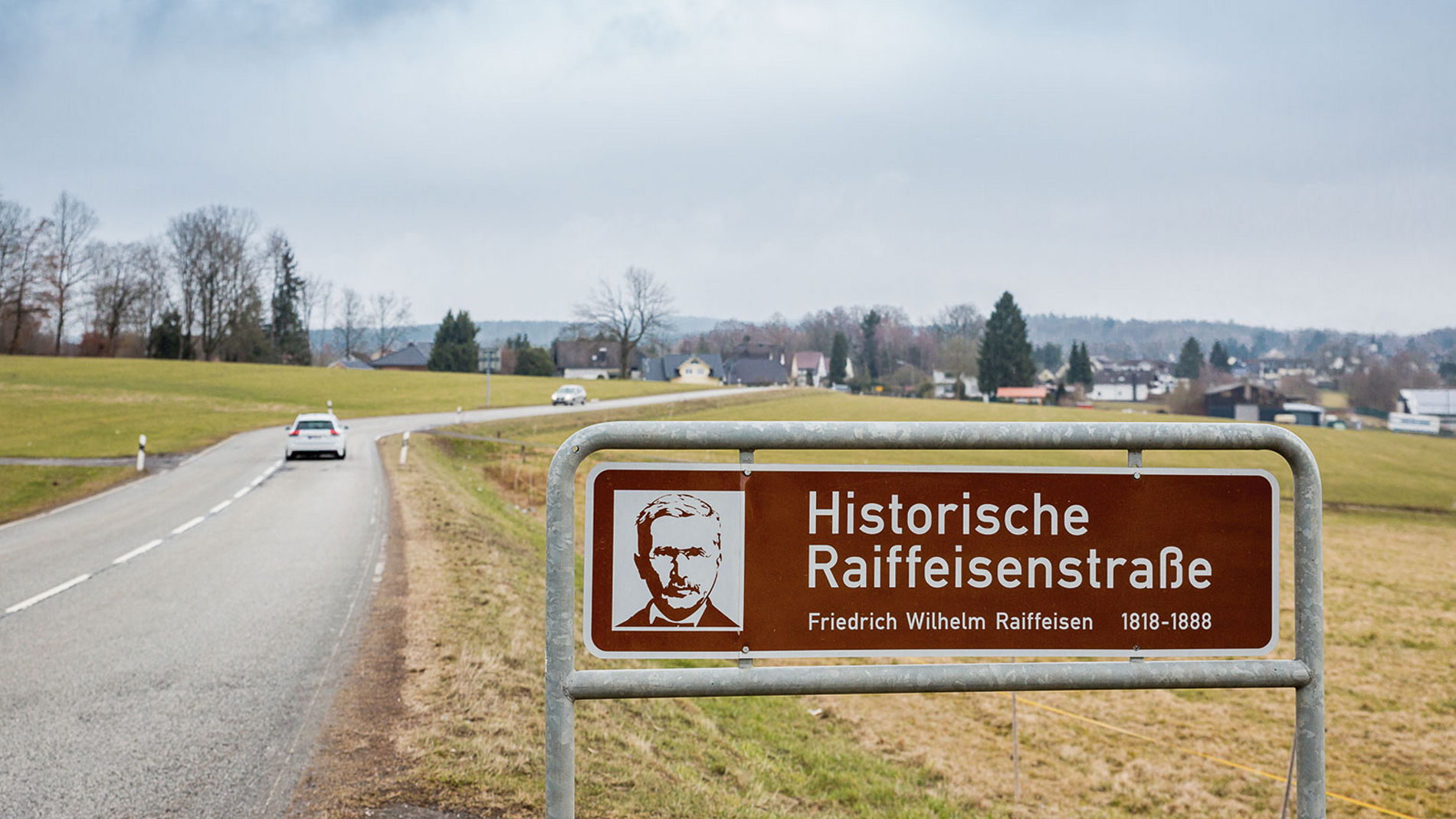 Das Bild zeigt ein Hinweisschild entlang einer Landstraße, das auf die "Historische Raiffeisenstraße" hinweist.
