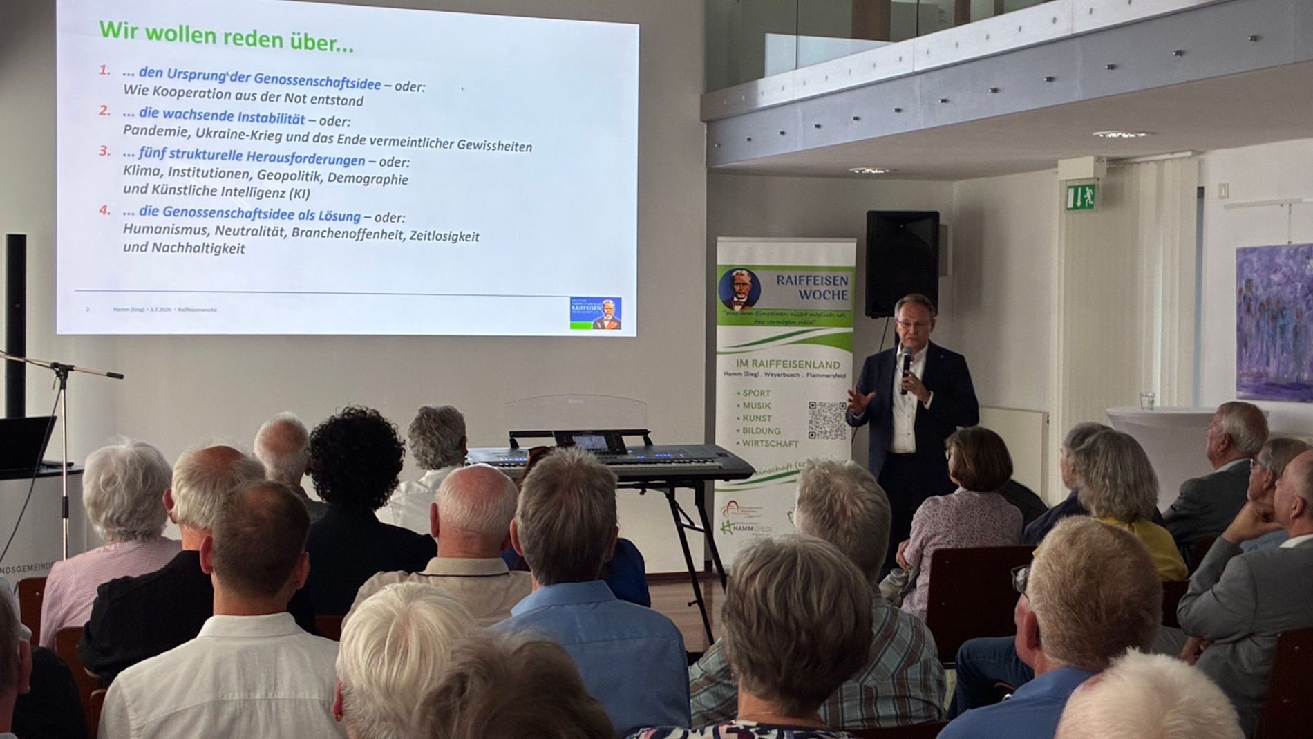 Eine Vortrags- oder Informationsveranstaltung in einem Innenraum: ein Redner steht vorne neben einer projizierten Präsentationsfolie mit dem Titel „Wir wollen reden über...“, davor sitzen mehrere Zuhörer in Reihen (überwiegend ältere Personen). Im Raum sind Tische, Stuhlreihen und eine Roll-up-Banner; es wirkt wie ein Gemeindesaal oder Veranstaltungsraum. (Ich kann keine Personen identifizieren.)