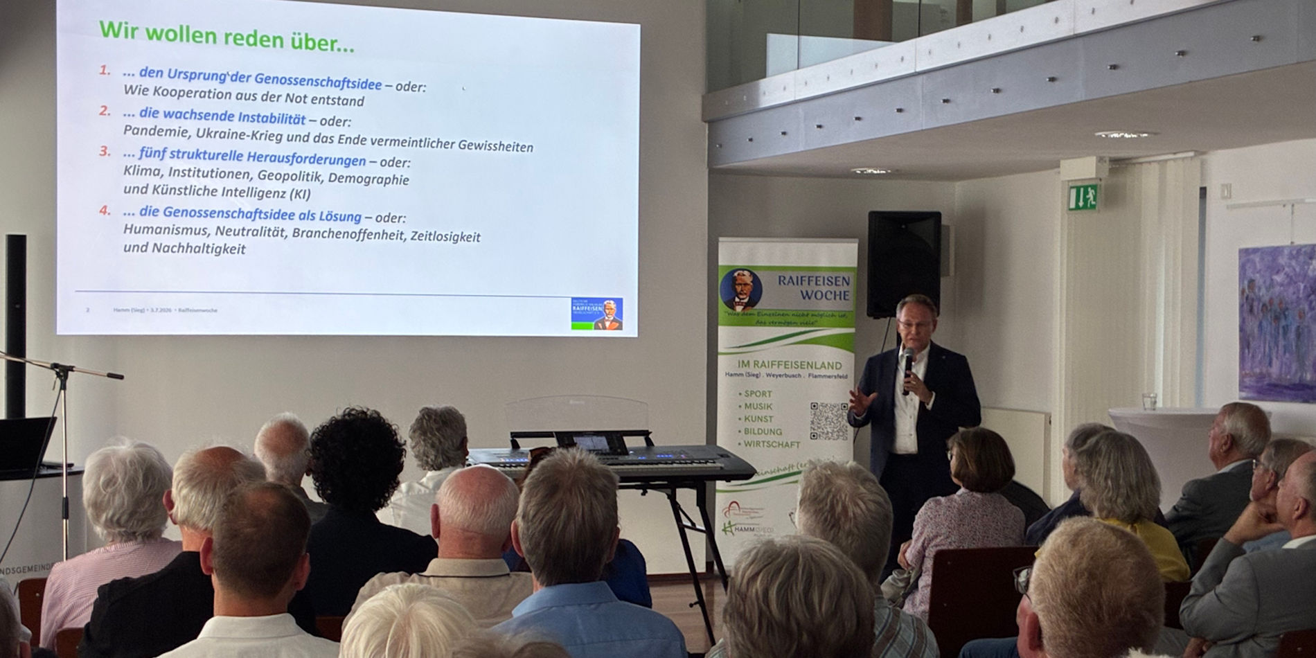 Eine Vortrags- oder Informationsveranstaltung in einem Innenraum: ein Redner steht vorne neben einer projizierten Präsentationsfolie mit dem Titel „Wir wollen reden über...“, davor sitzen mehrere Zuhörer in Reihen (überwiegend ältere Personen). Im Raum sind Tische, Stuhlreihen und eine Roll-up-Banner; es wirkt wie ein Gemeindesaal oder Veranstaltungsraum. (Ich kann keine Personen identifizieren.)