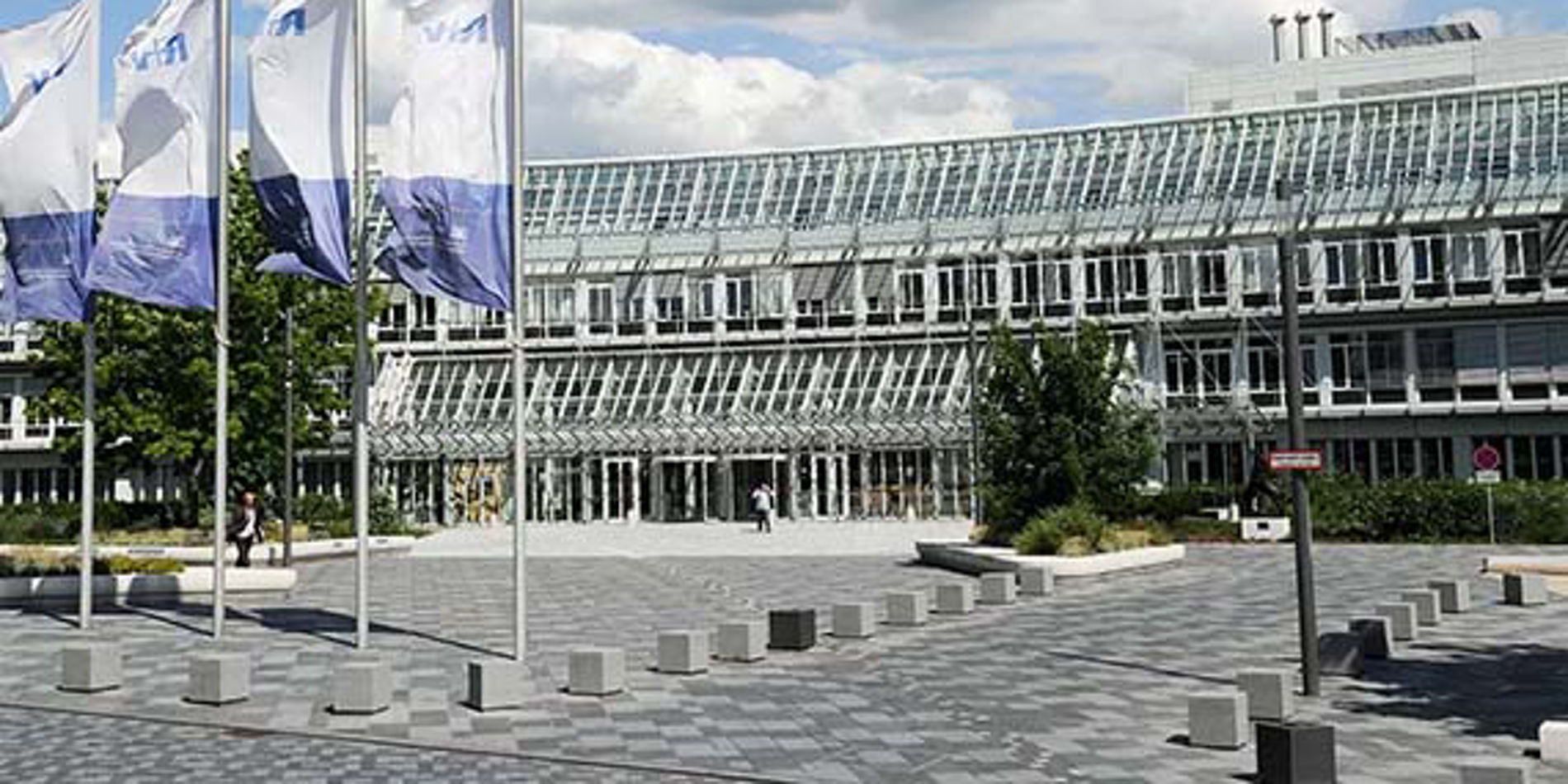 Ein modernes Gebäude mit großer Glasfassade (Büro- oder Verwaltungsgebäude) an einem gepflasterten Vorplatz. Im Vordergrund sind mehrere Fahnenmasten mit wehenden Fahnen, quadratische Poller und Sitzblöcke auf dem Platz. Im Hintergrund Bäume und ein teils bewölkter Himmel.