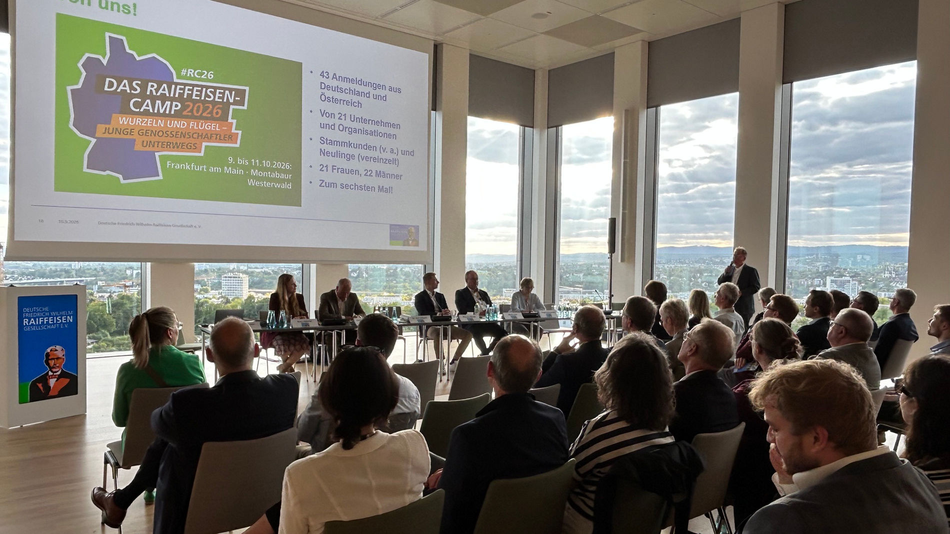 Eine Konferenz-/Präsentationsszene in einem hellen Tagungsraum: mehrere Reihen sitzender Teilnehmender schauen auf ein großes Leinwand-Display mit einer bunten Präsentationsfolie („Das Raiffeisen Camp 2025“ erkennbar). Vorne sitzt ein Podium/Tisch mit mehreren Personen (Podiumsteilnehmer/Panel), rechts steht eine Person, die vermutlich spricht. Große Fenster bieten Ausblick auf die Stadt und den Himmel.