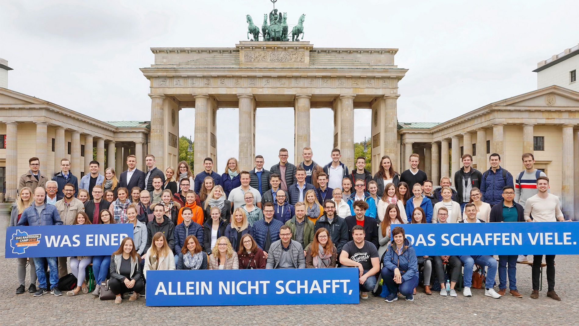 Personengruppe vor dem Brandenburger Tor in Berlin, überwiegend junge Menschen. Sie halten ein Banner: Was einer allein nicht shcafft, das schaffen viele.