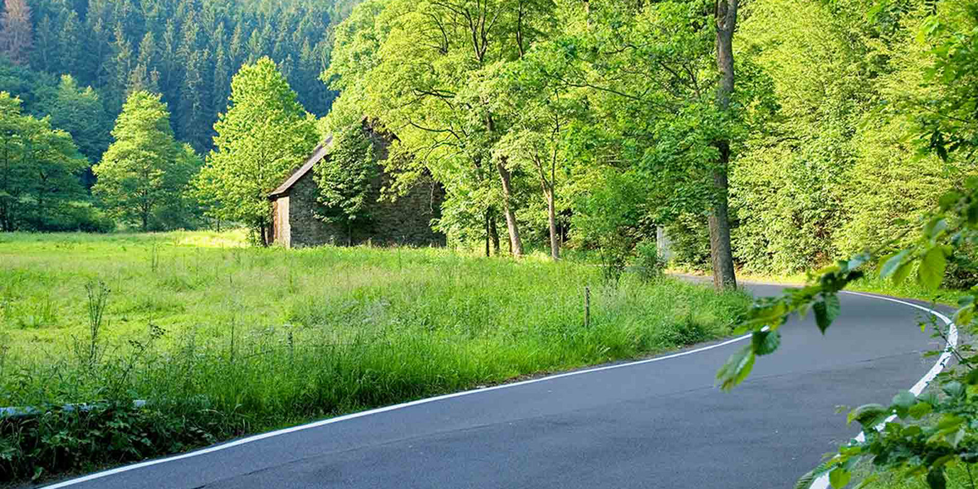 Das Bild stellt eine malerische ländliche Szene dar, in der eine kurvige Landstraße durch eine friedliche, grüne Umgebung mit üppigen Bäumen und Gras verläuft. Rechts und links der Straße befindet sich dichtes Blattwerk, das den Eindruck von natürlicher Abgeschiedenheit verleiht. Im Hintergrund ist ein kleines, rustikales Gebäude sichtbar, vermutlich ein alter Bauernhof oder eine Scheune, das sich harmonisch in die grüne Landschaft einfügt. Die Szenerie wirkt ruhig und entspannend, und die sonnige Beleuchtung lässt die Farben intensiver erscheinen.