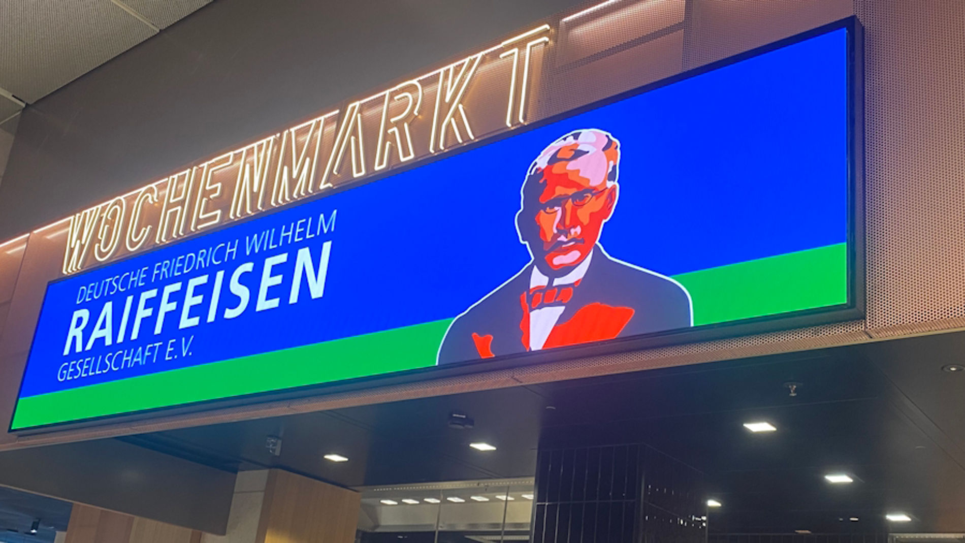 Ein beleuchtetes Werbeschild/Leuchtkasten in einer Innenhalle (vermutlich eine Bankfiliale oder ein Kassenbereich). Auf dem Schild steht groß „HOCHMARKT“ und darunter „RAIFFEISEN“ sowie weitere Textzeilen. Dazu ist ein stilisiertes Porträt eines Mannes in Anzug zu sehen. Im Vordergrund sind Schalter/Arbeitsplätze und Personen erkennbar.