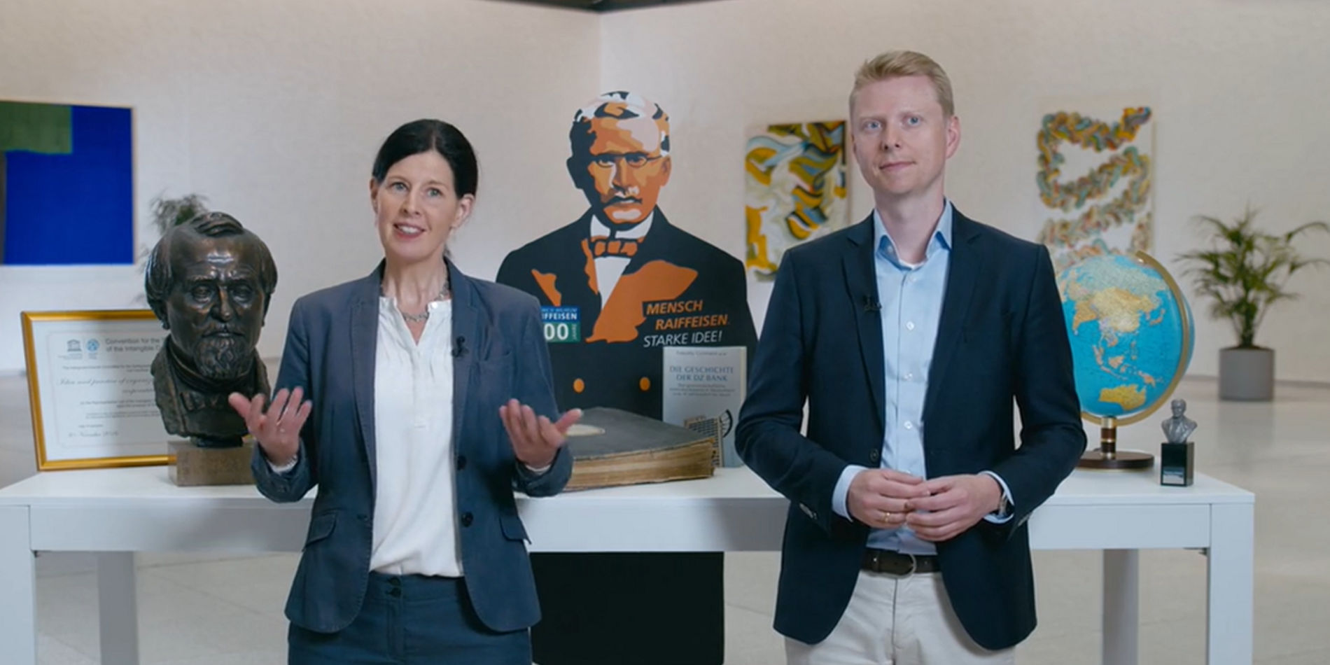 Das Bild zeigt eine Umgebung, die ausgestellte Gegenstände präsentiert. Rechts befindet sich ein Globus auf einem Ständer. Zentral ist eine stilisierte Figur in einer Uniform zu sehen mit einem Schild, das möglicherweise der Name eines Teams oder einer Organisation ist. Links steht ein gerahmtes Zertifikat oder Plakat und eine Skulptur oder Büste. Pflanzen sind auch im Hintergrund zu sehen.