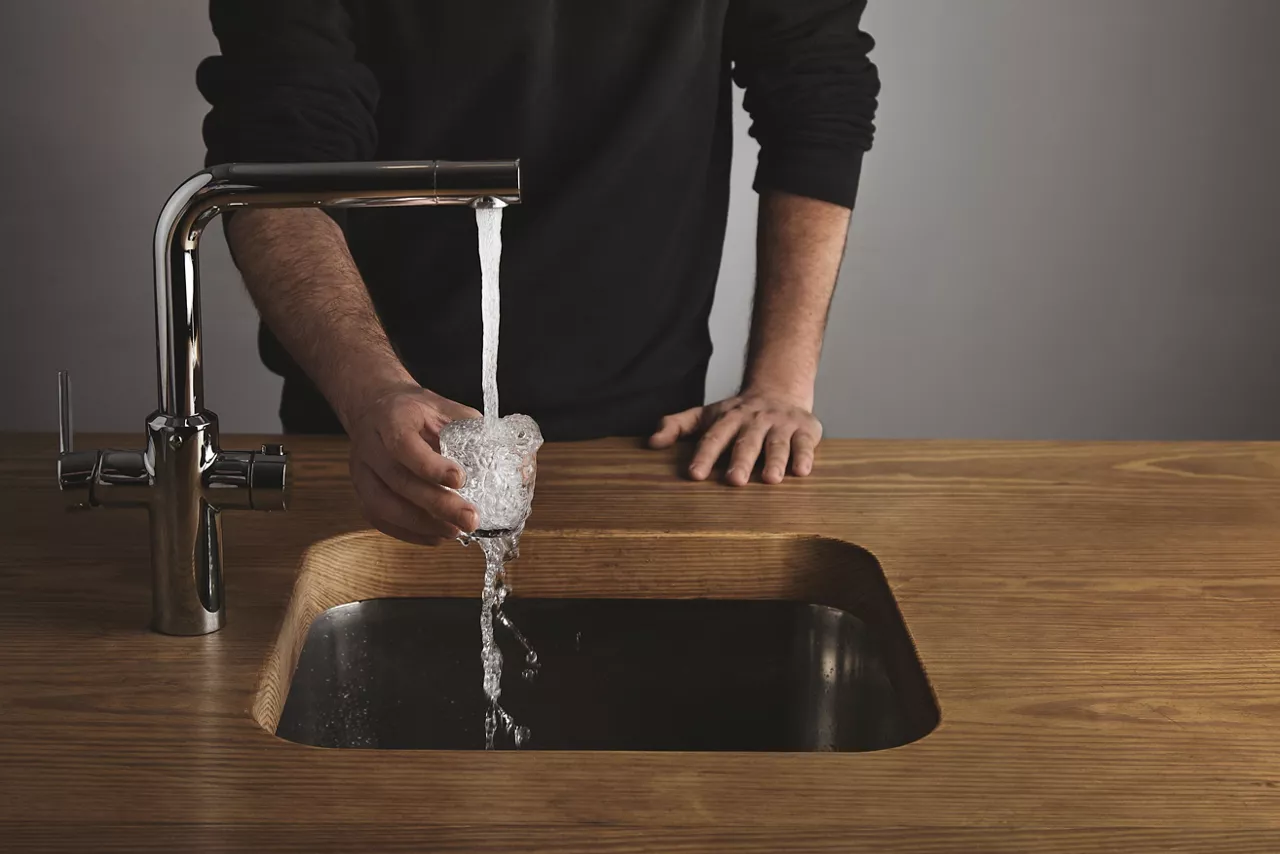 Brutal barista in black sweatshot behind thick wooden table rinses small tranparent glass with water under silver metal tap in cafe shop. Water drops out of glass.