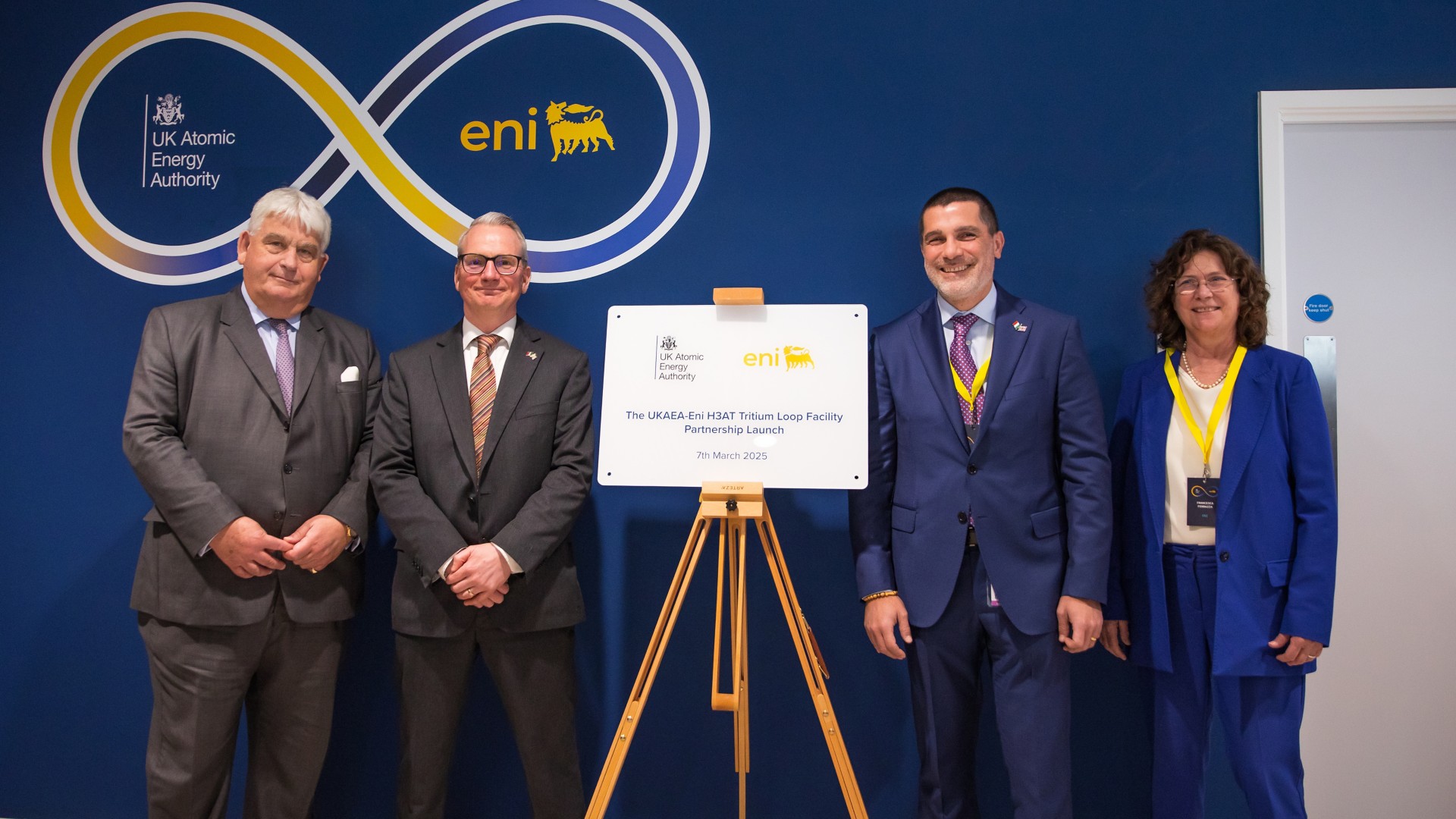 three men and a woman smile as they pose near a sign announcing the partnership
