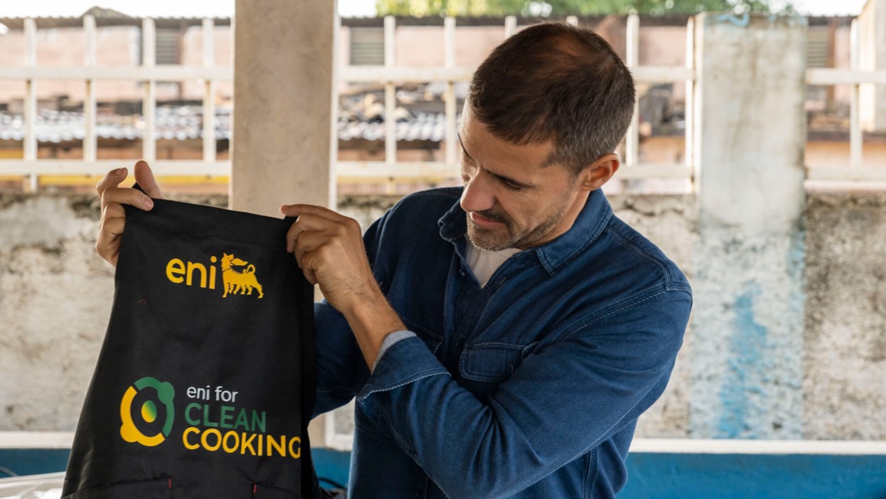 Man displaying a black kitchen apron with a yellow Eni logo and the words 'Clean Cooking'