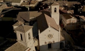 Veduta aerea della Basilica di San Benedetto a Norcia, con la facciata in pietra chiara decorata da un grande rosone centrale e da tre portali gotici. La chiesa è circondata dai tetti in laterizio del centro storico, illuminati dalla luce calda del tramonto.