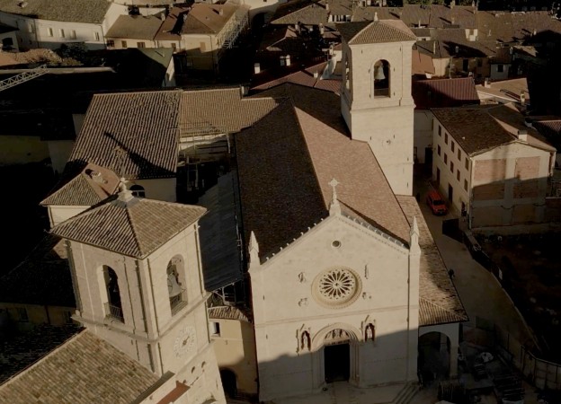 Aerial view of the Basilica of Saint Benedict in Norcia, featuring a light-stone façade with a large rose window and three Gothic portals, surrounded by the terracotta rooftops of the historic center bathed in warm sunset light.