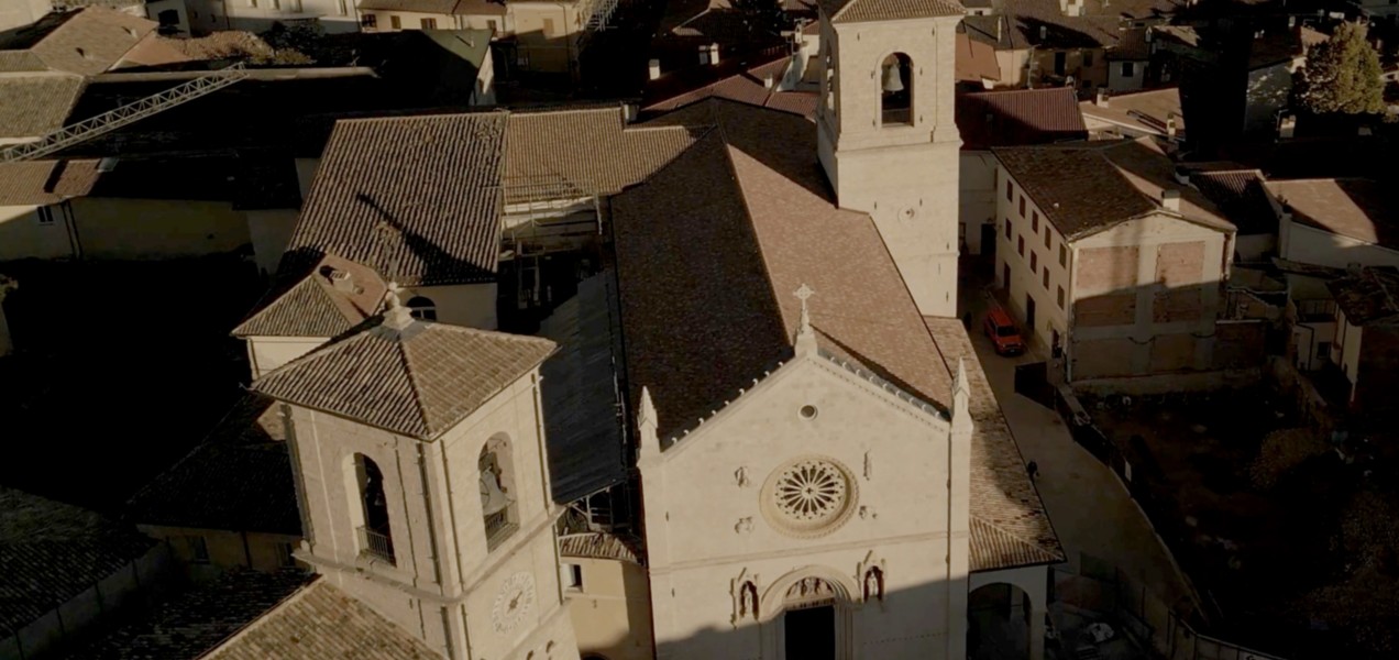 Veduta aerea della Basilica di San Benedetto a Norcia, con la facciata in pietra chiara decorata da un grande rosone centrale e da tre portali gotici. La chiesa è circondata dai tetti in laterizio del centro storico, illuminati dalla luce calda del tramonto.