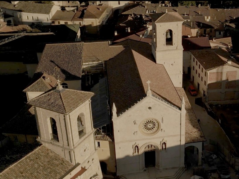 Aerial view of the Basilica of Saint Benedict in Norcia, featuring a light-stone façade with a large rose window and three Gothic portals, surrounded by the terracotta rooftops of the historic center bathed in warm sunset light.