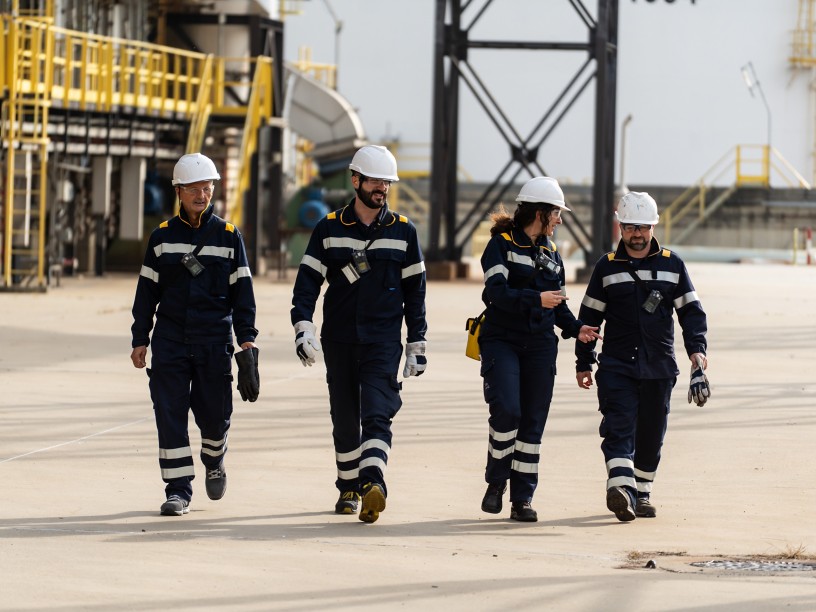 Quattro lavoratori e lavoratrici con caschi di sicurezza e tute da lavoro camminano insieme all’interno di un impianto industriale, conversando tra loro, con strutture e tubazioni sullo sfondo