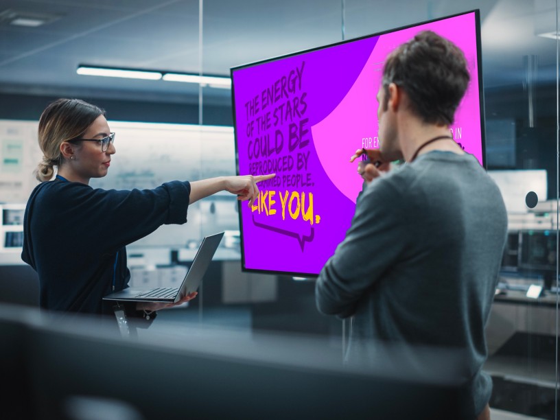 A young man and a young woman look at a computer monitor displaying a motivational quote inside an office