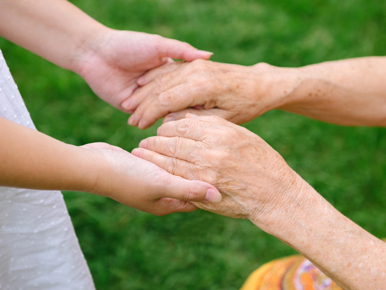 Le mani di una persona giovane sorreggono con delicatezza le mani di una persona anziana, con uno sfondo verde sfocato.