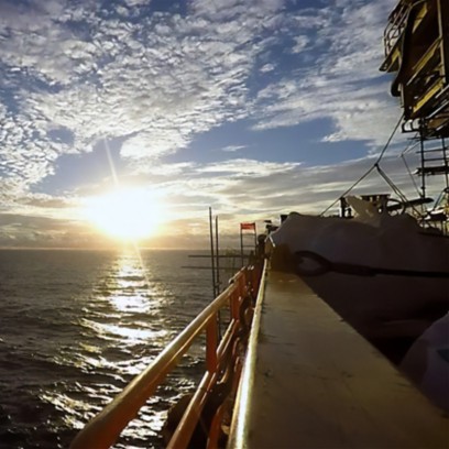 Sunset view of the sea from the deck of an offshore vessel, with the sun low on the horizon and golden reflections on the water