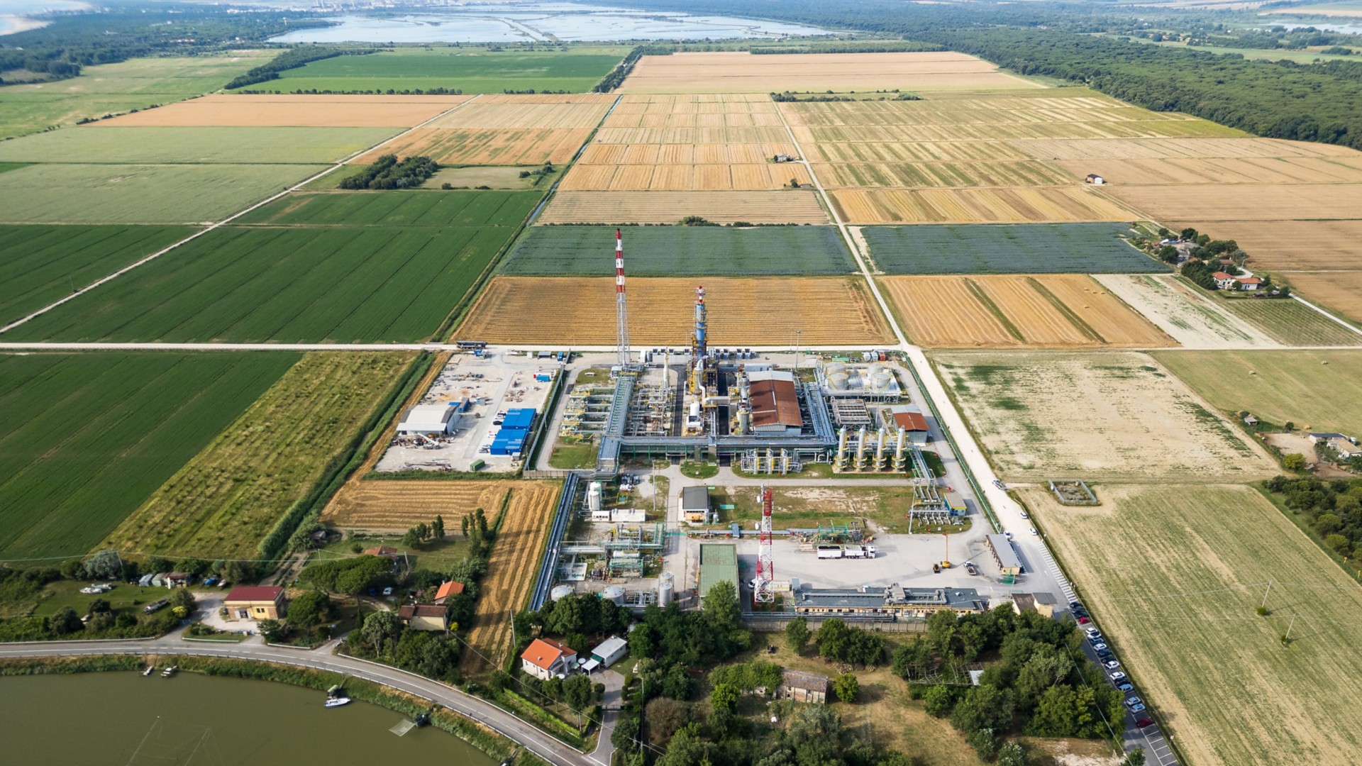 Veduta aerea di un impianto industriale circondato da campi agricoli nella campagna. In primo piano un fiume con una strada che lo costeggia, sullo sfondo campi coltivati e una zona boschiva.