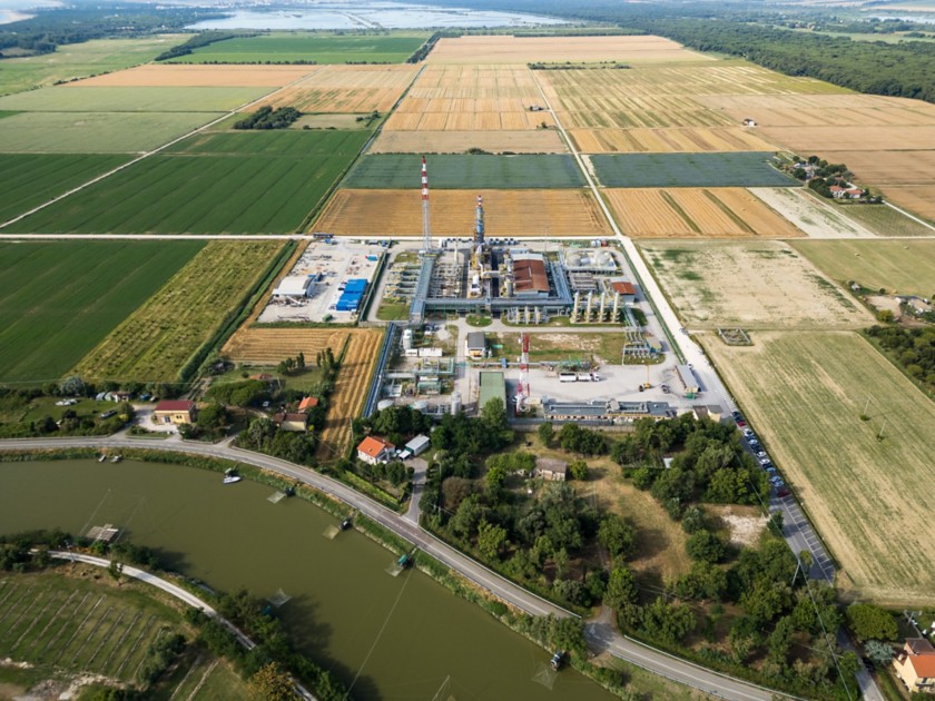 Veduta aerea di un impianto industriale circondato da campi agricoli nella campagna. In primo piano un fiume con una strada che lo costeggia, sullo sfondo campi coltivati e una zona boschiva.