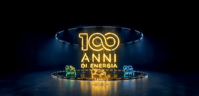 Scritta luminosa “100 anni di energia” in neon giallo al centro di uno spazio buio, con il Cane a sei zampe illuminato 