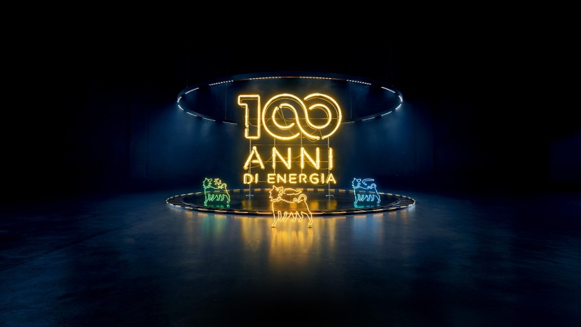 Scritta luminosa “100 anni di energia” in neon giallo al centro di uno spazio buio, con il Cane a sei zampe illuminato 