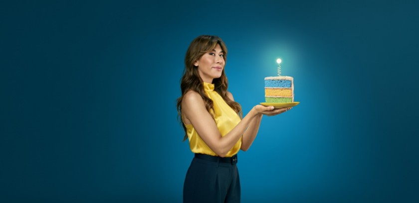 Virginia Raffaele con un top giallo tiene una fetta di torta a strati colorati, sormontata da una lampadina accesa, su sfondo blu petrolio.