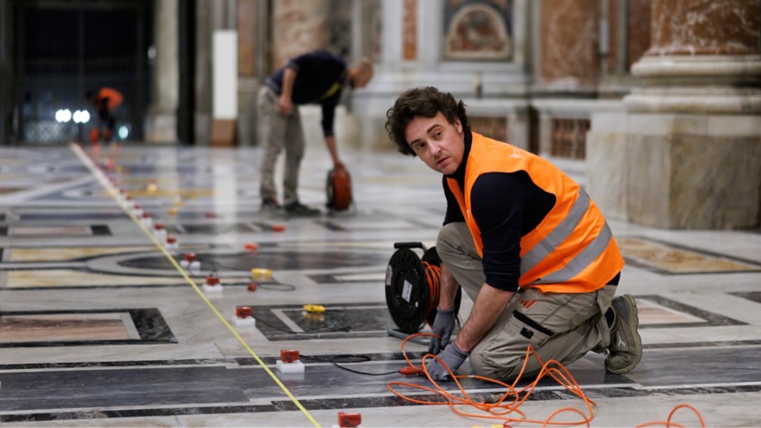 Primo piano di tecnici che stendono cavi e posizionano sensori di precisione sul pavimento in marmo della Basilica 