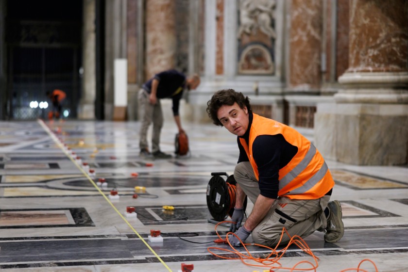 Primo piano di tecnici che stendono cavi e posizionano sensori di precisione sul pavimento in marmo della Basilica 