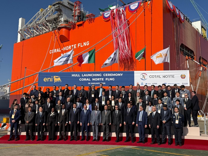 Group photo of authorities, executives and industrial partners at the Coral Norte FLNG hull launching ceremony, standing in front of the orange facility structure with the official Eni banner and international flags.