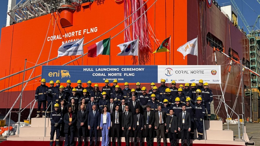 Group photo from the Coral Norte FLNG hull launching ceremony: executives and workers standing in front of the large orange industrial structure, with the official event banner and international flags displayed.