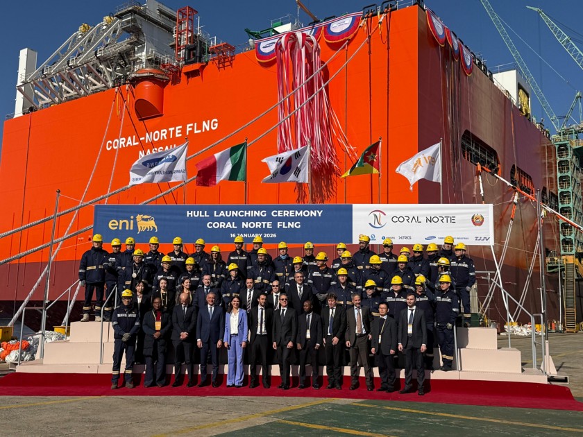 Foto di gruppo durante la cerimonia di varo dello scafo Coral Norte FLNG: dirigenti e lavoratori in primo piano davanti alla grande struttura industriale arancione dell’impianto, con striscione ufficiale dell’evento e bandiere internazionali.