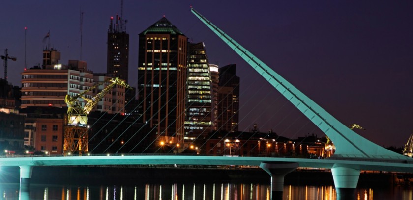 Night skyline of Buenos Aires featuring the illuminated Puente de la Mujer, city lights reflected on the water, and modern buildings in the Puerto Madero district.