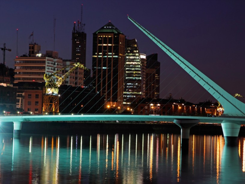Night skyline of Buenos Aires featuring the illuminated Puente de la Mujer, city lights reflected on the water, and modern buildings in the Puerto Madero district.