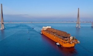 Cargo ship passes under a cable-stayed bridge, escorted by tugboats beneath a clear sky.
