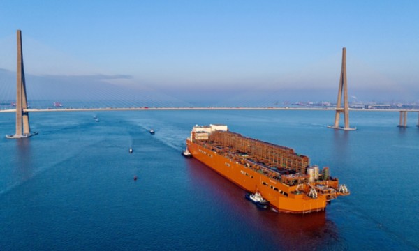 Nave cargo attraversa un ponte strallato, scortata da rimorchiatori sotto cielo sereno.