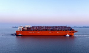 Orange cargo ship escorted by two tugboats in a harbor at sunset.